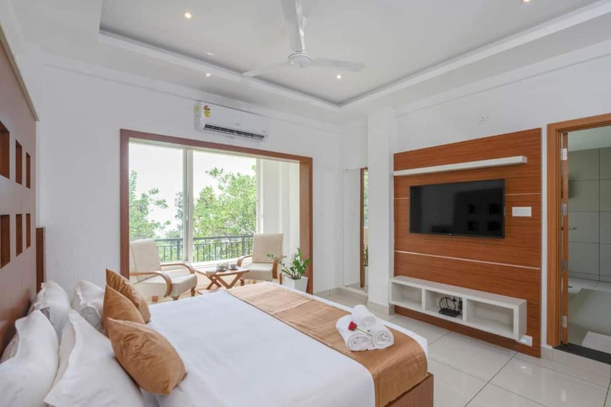 A modern hotel room with a large bed, a TV mounted on the wall, and a balcony with a view of greenery.