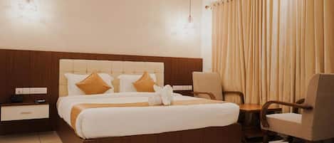 Habitación de hotel con una cama grande, cabecera de madera, mesita de noche, una silla y ventana con cortinas.