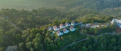 An aerial view of a residential area with houses surrounded by dense forest, a winding road, and a large building with multiple floors.