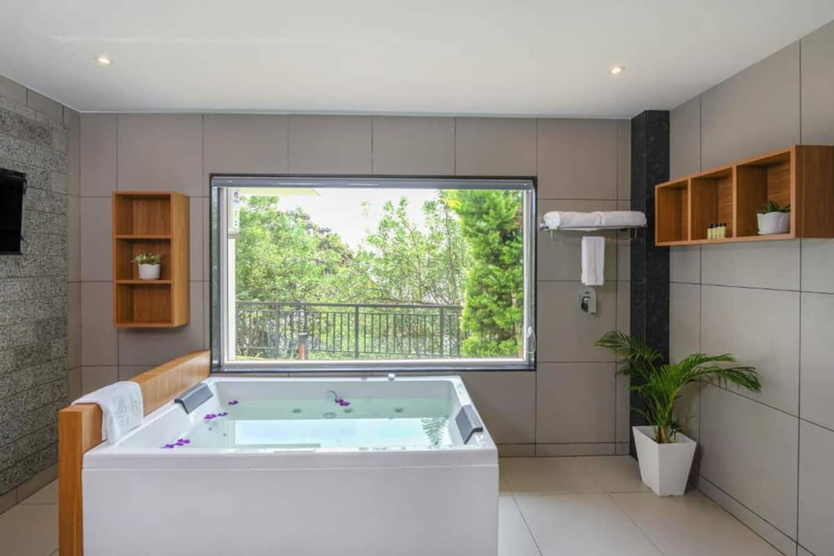 A modern bathroom with a large bathtub, a window with a view of trees, and wooden shelves with toiletries.