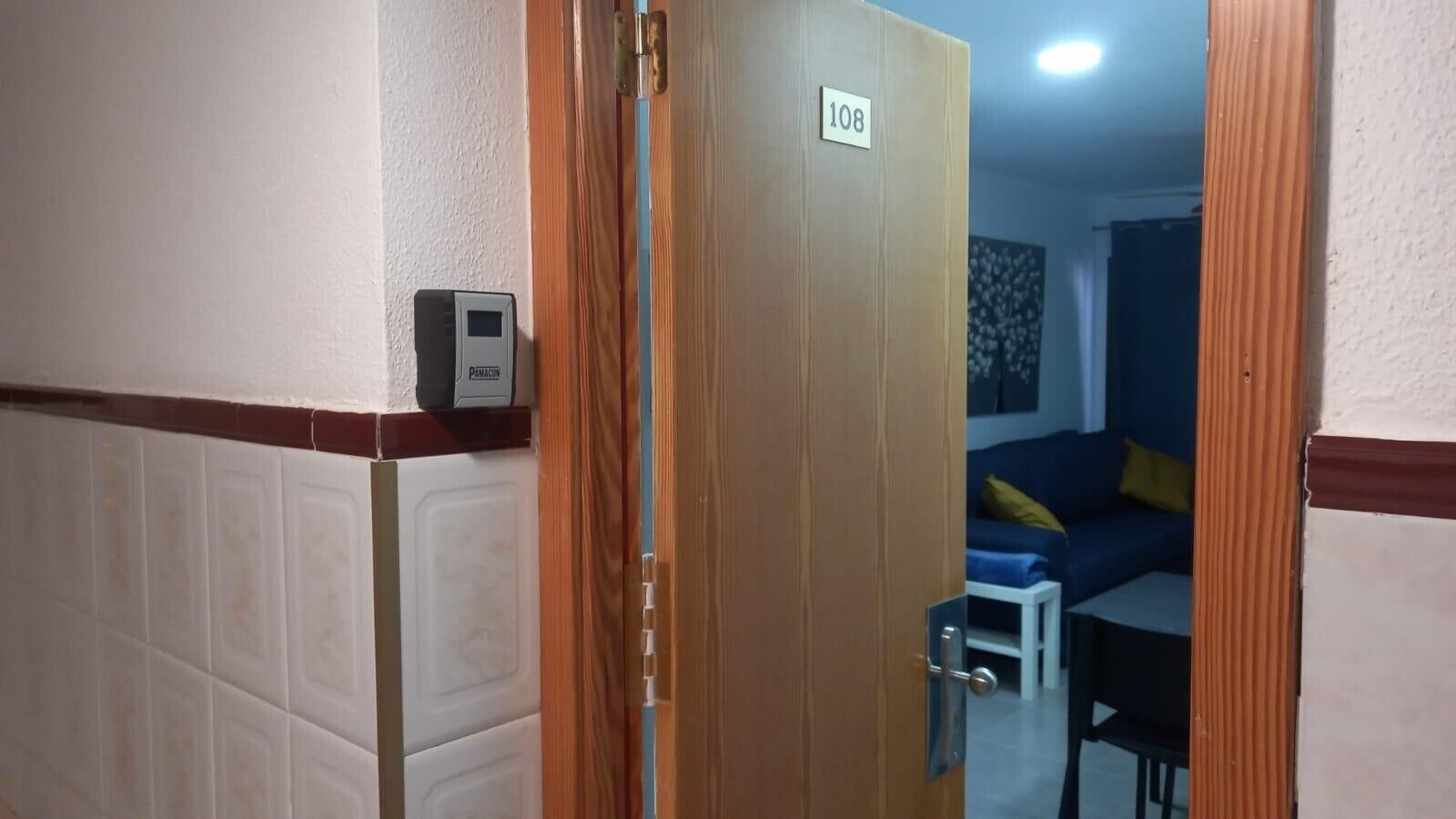 Ein Korridor mit einer Holztür mit der Aufschrift '108', einer gefliesten Wand und einem Blick in einen Wohnbereich mit Sofa und kleinem Tisch.