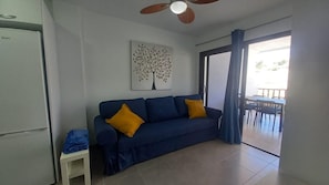 A living room with a blue sofa, a ceiling fan, a white refrigerator, and a dining area with chairs and a table.