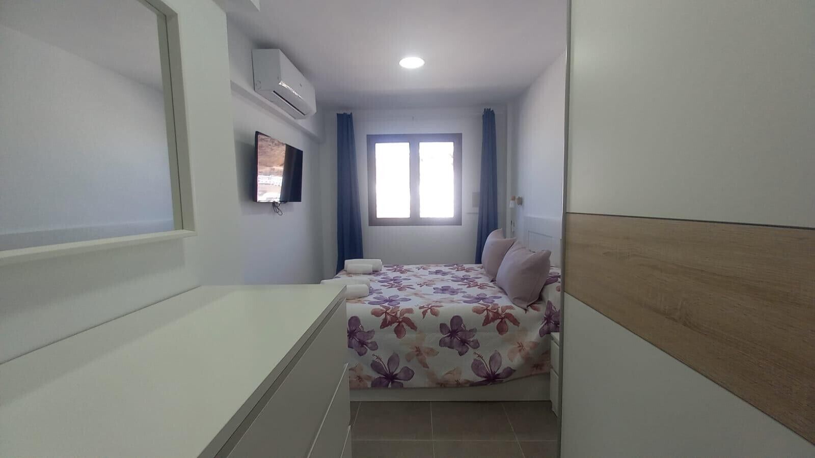 A bedroom with a bed, a TV, an air conditioning unit, and a window.
