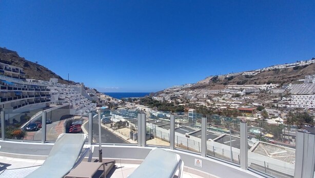 A high vantage point overlooking a coastal city with modern architecture, a clear blue sky, and a view of the sea.