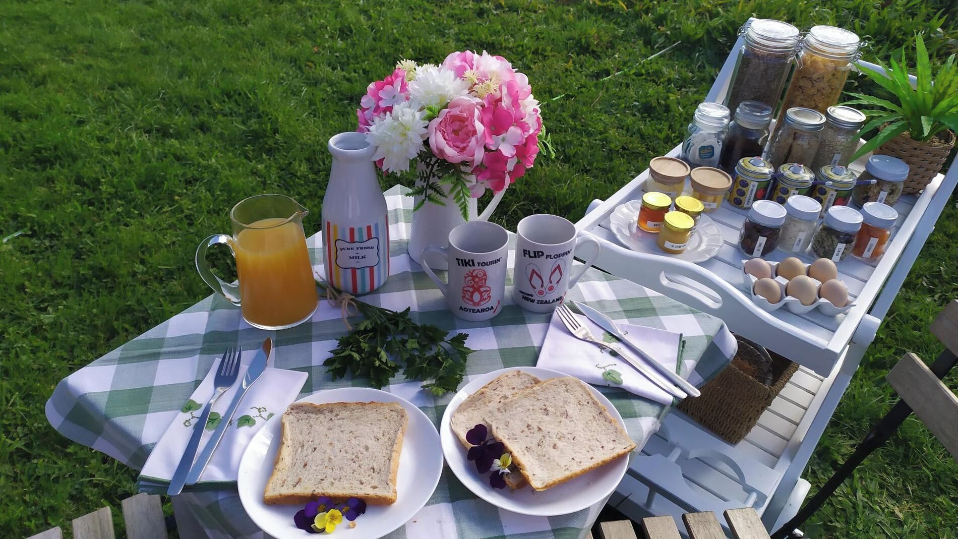 A picnic setup with a checkered tablecloth, two plates of bread, a pitcher of juice, a vase of flowers, and a tray of condiments and eggs.