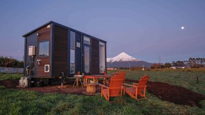 A modern cabin with a dining area, outdoor seating, and a view of a snow-capped mountain. - Taranaki Creekside TUI by Tiny Away (Midhirst)