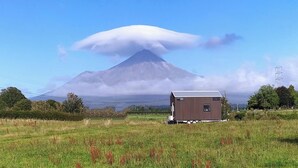 A small wooden cabin with a metal roof in a grassy field, with a mountain and clouds in the background. - Taranaki Creekside TUI by Tiny Away (Midhirst)