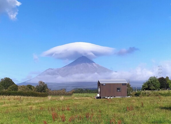 A small wooden cabin with a metal roof in a grassy field, with a mountain and clouds in the background. - Taranaki Creekside TUI by Tiny Away (Midhirst)