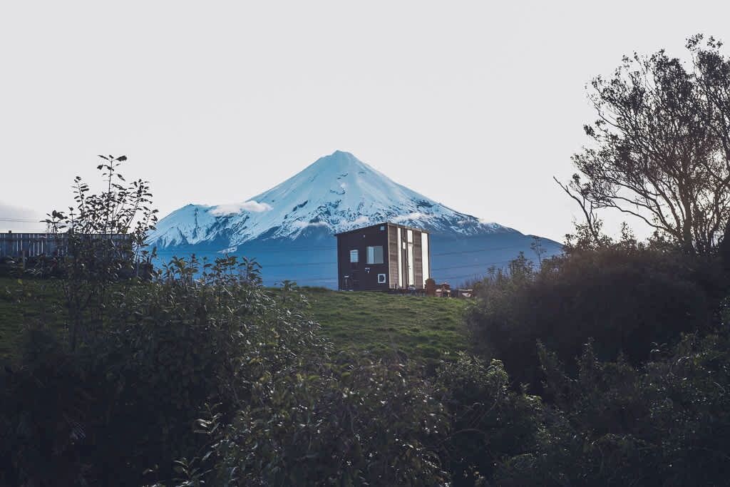 Un edificio pequeño y moderno con techo plano y ventanas amplias, ubicado en un área con césped y una montaña nevada al fondo.