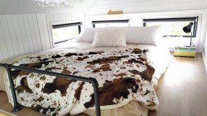 A bedroom with a bed, a white pillow, a cow print blanket, a bedside lamp, and a stack of books. - Taranaki Creekside TUI by Tiny Away (Midhirst)