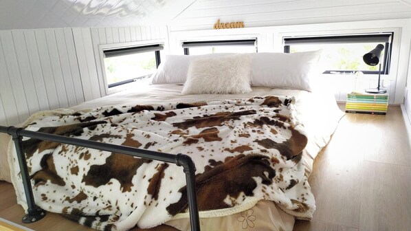 A bedroom with a bed, a white pillow, a cow print blanket, a bedside lamp, and a stack of books. - Taranaki Creekside TUI by Tiny Away (Midhirst)