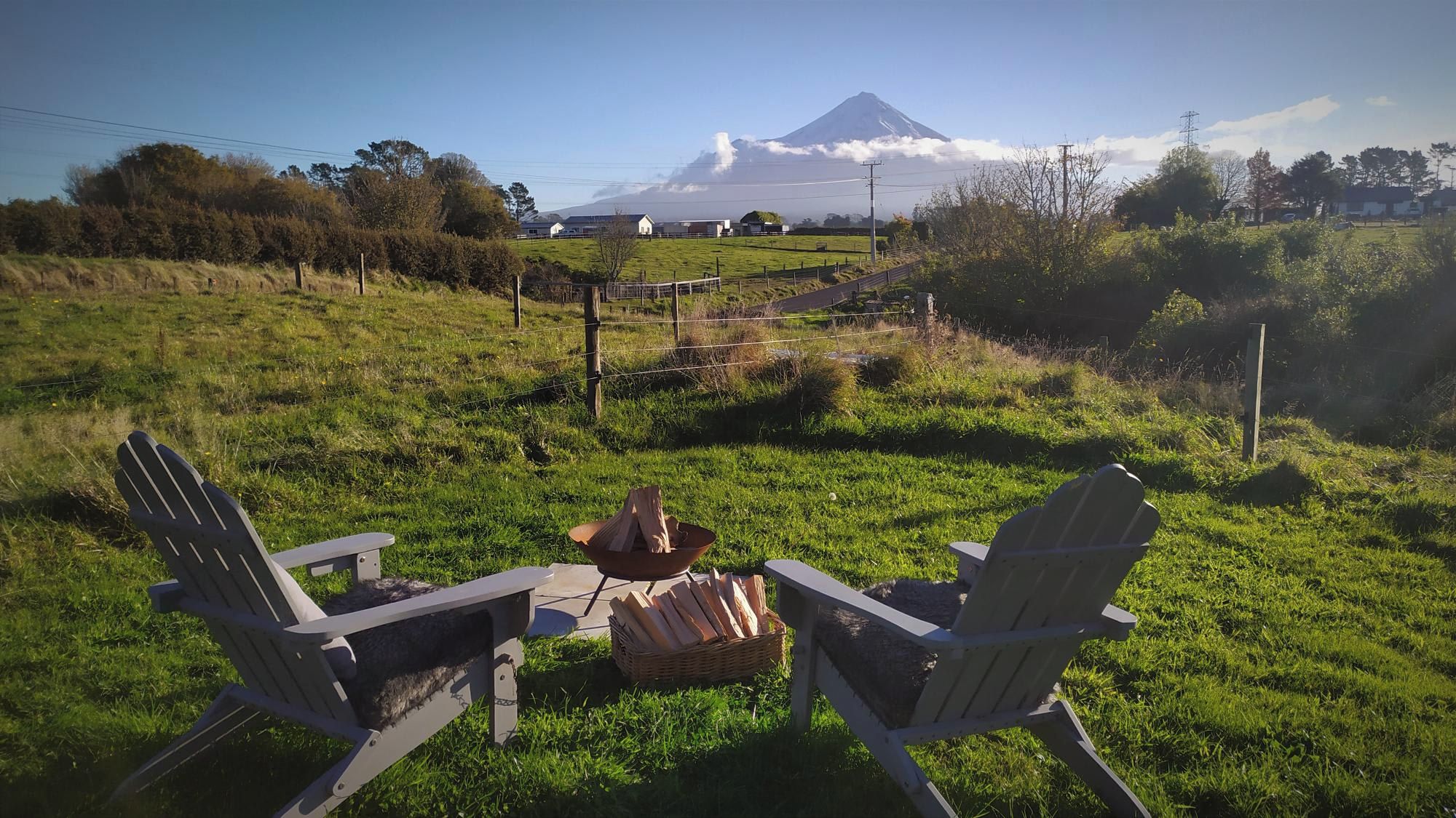 Two Adirondack chairs facing each other with a fire pit in between, set on a grassy field with a mountain in the background.