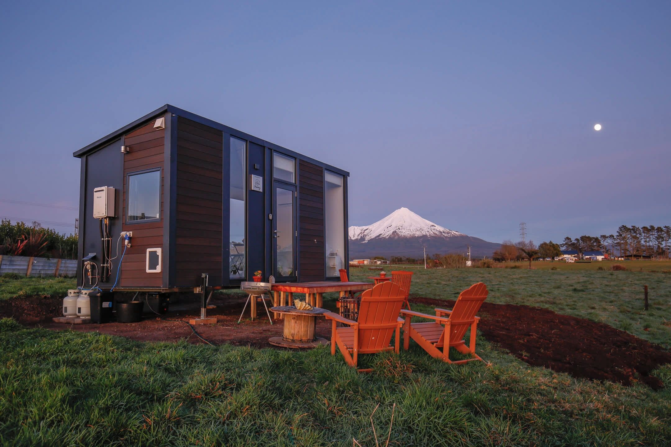 A modern cabin with a dining area, outdoor seating, and a view of a snow-capped mountain.