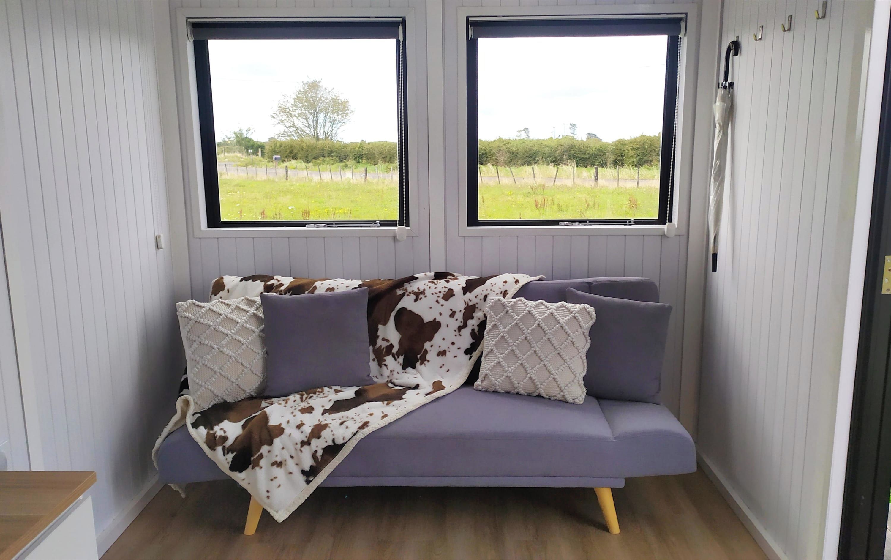 A room with a purple sofa, two windows with a view of a grassy field, and a white wall with a door.