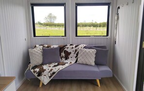 Ein Zimmer mit einem violetten Sofa, zwei Fenstern mit Blick auf ein grünes Feld und einer weißen Wand mit einer Tür.