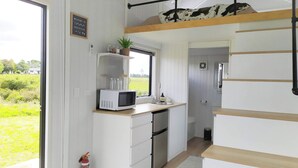 A compact kitchen with a microwave, sink, and a small refrigerator. There's a window with a view of greenery outside. - Taranaki Creekside TUI by Tiny Away (Midhirst)