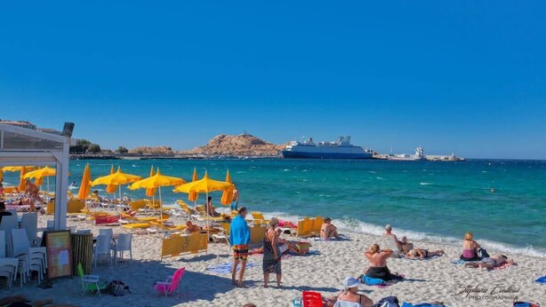 Een strand met gele parasols en ligbedden, een schip op de achtergrond en een rotsachtige richel.