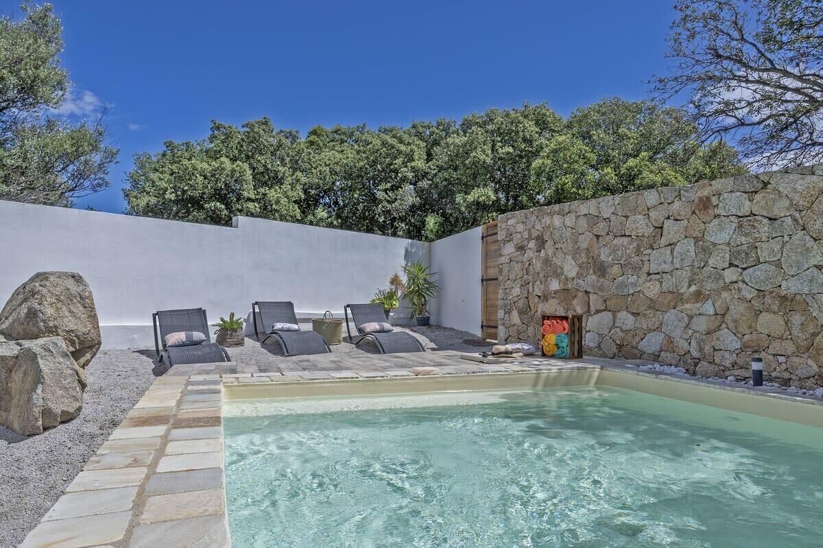 A pool area with lounge chairs, a stone wall, and a view of trees.