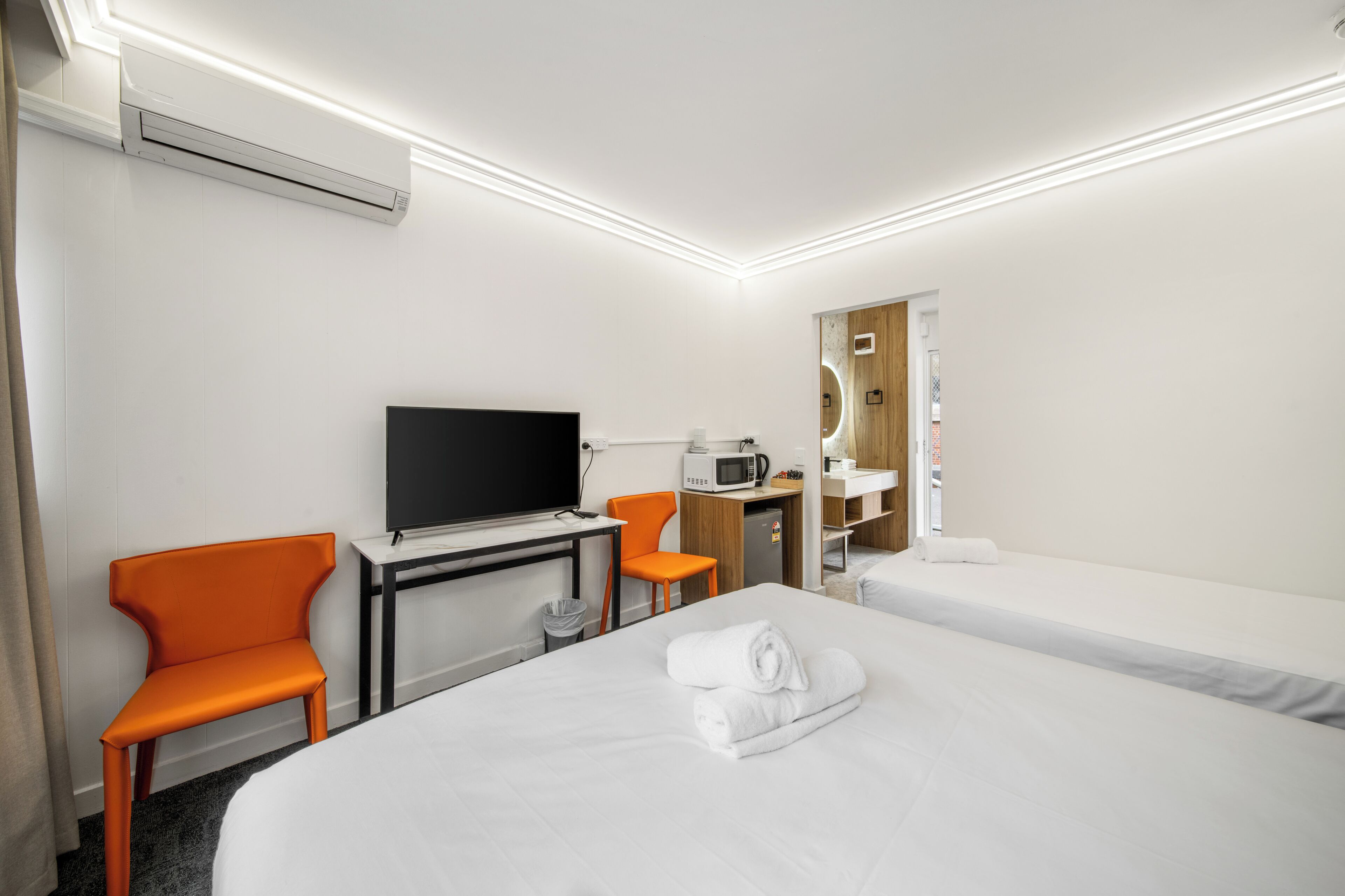 Quarto de hotel com cama, escrivaninha com TV de tela plana, cadeira laranja, micro-ondas e pequena cozinha equipada.
