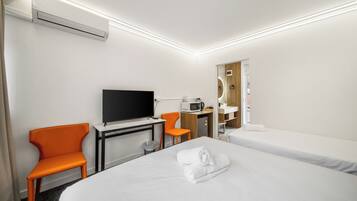 Quarto de hotel com cama, escrivaninha com TV de tela plana, cadeira laranja, micro-ondas e pequena cozinha equipada.