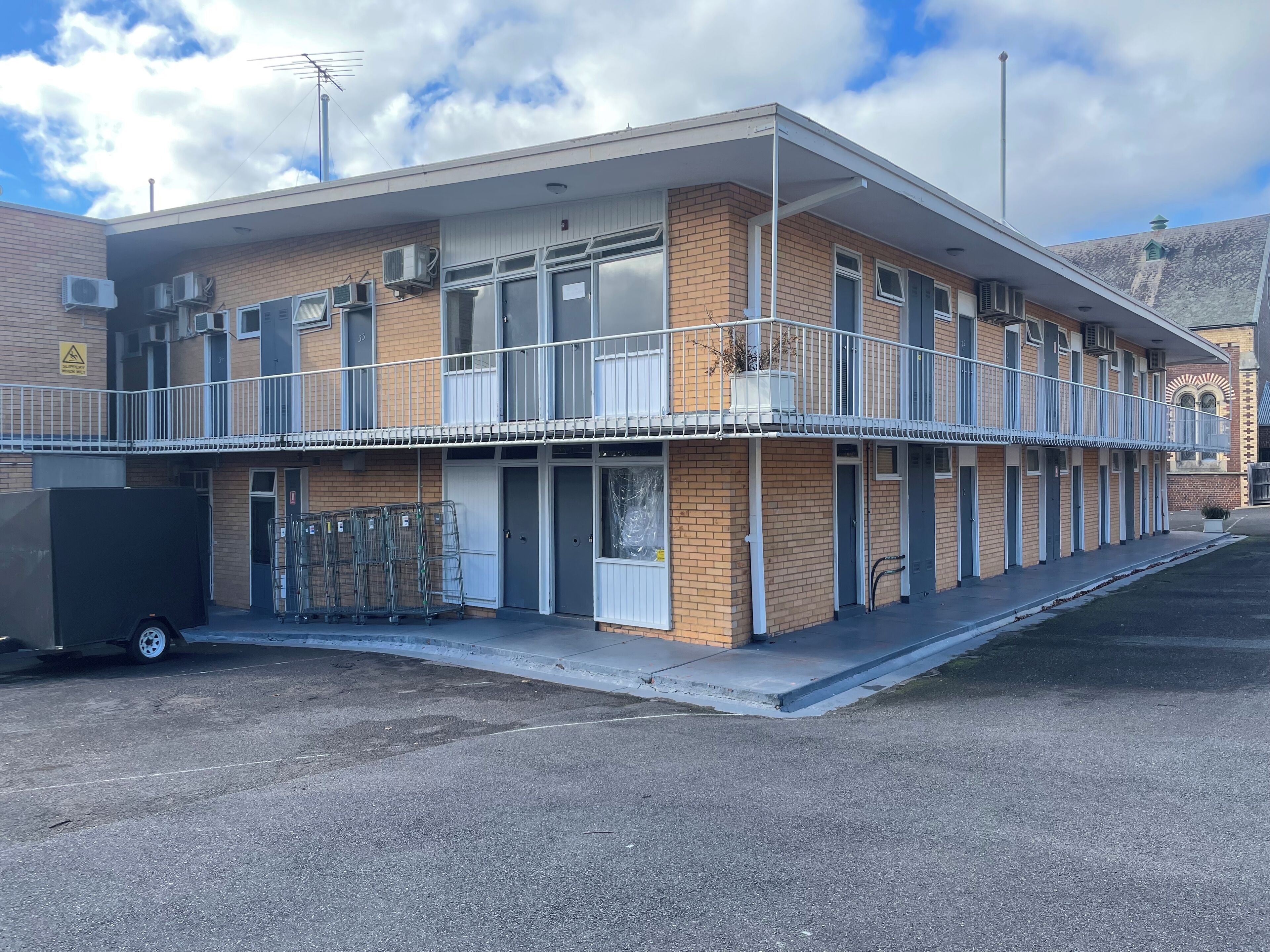 A two-story motel with a flat roof, multiple air conditioning units, and a parking area with a trailer.