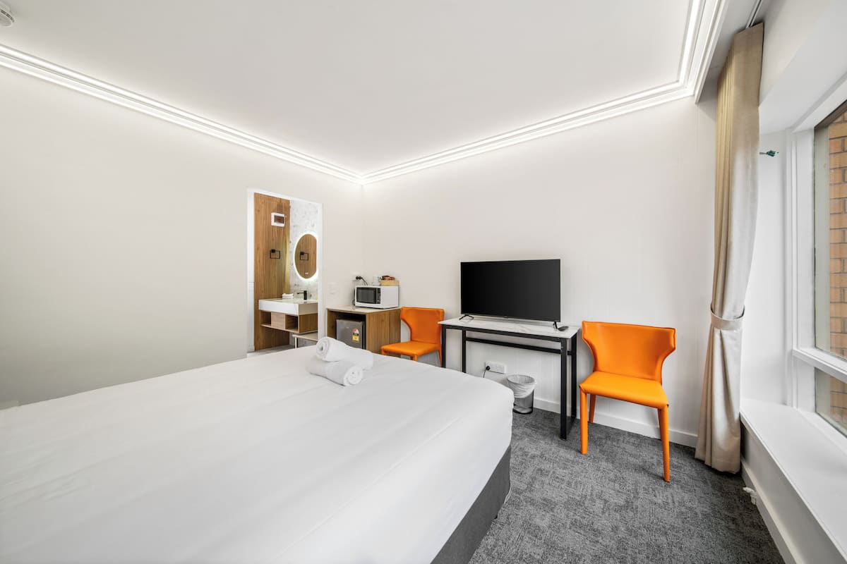 A hotel room with a large bed, a desk with a flat-screen TV, an orange chair, a microwave, and a small trash bin.