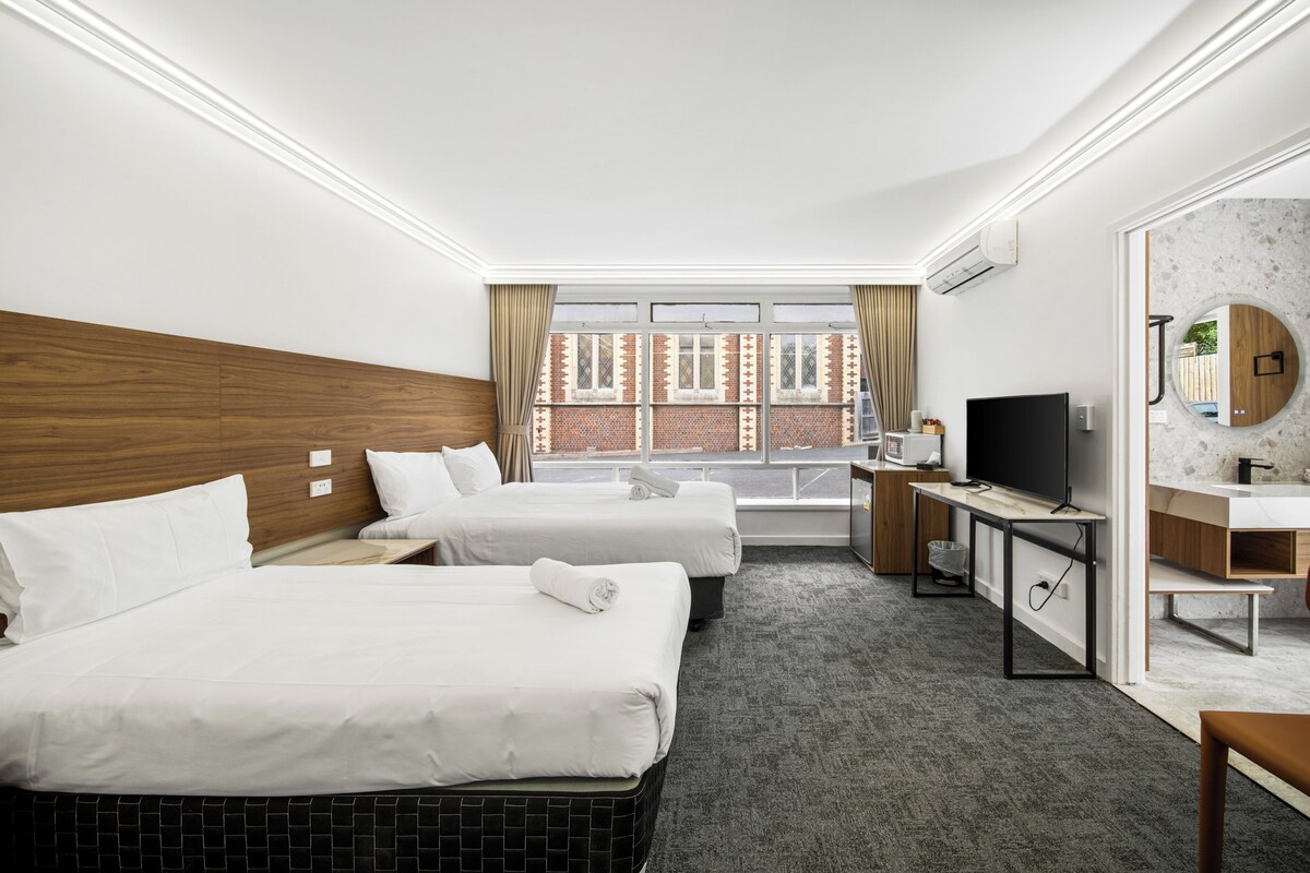 A hotel room with two beds, a desk, a TV, and a bathroom with a mirror and sink.