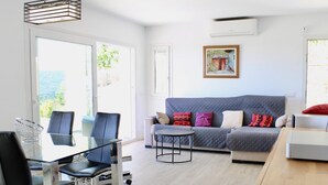A modern living room with a grey sofa, a glass dining table, and black chairs. There is a painting on the wall and a window with a view of greenery. - House with garden and private pool 6km beach (Calonge)