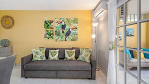 Un soggiorno con un divano grigio, opere d'arte a tema tropicale, un oggetto decorativo rotondo e una finestra vista.
