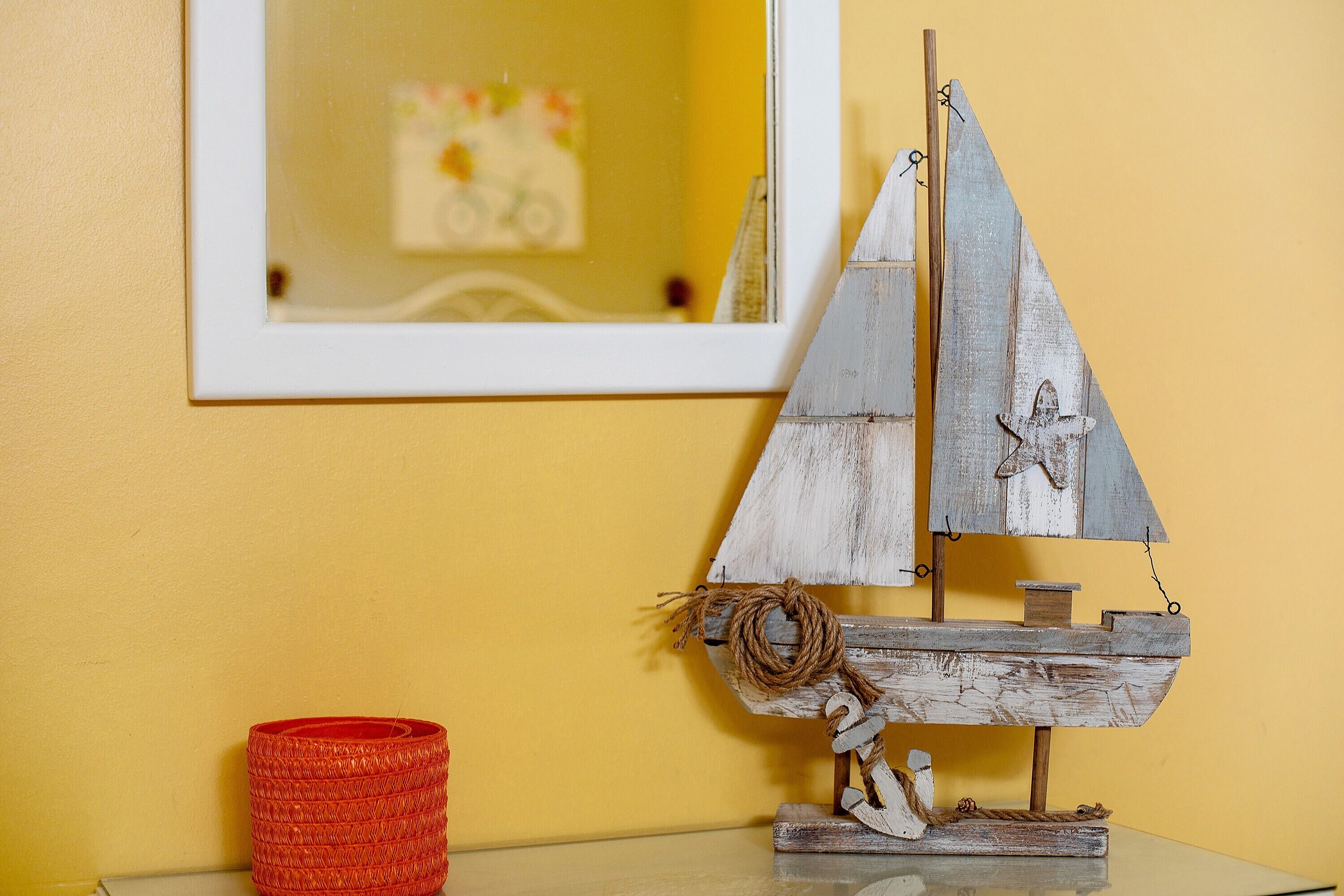 A model sailboat on a table with a yellow wall and a framed picture in the background.