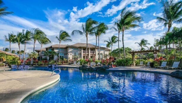 Een zwembadgedeelte van een resort, met palmbomen, ligbedden en een gebouw op de achtergrond.