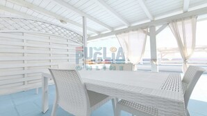 Un espace extérieur abrité, doté d'une table et de chaises en osier blanc, offrant une vue sur la mer et surmonté d'une structure au plafond également blanche.