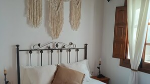 A bed with white bedding, a wooden headboard, macrame wall hangings, a wooden nightstand with a candle, and a window with curtains. - Holiday Home 'Casa Rural Villa Campito' with Mountain View, Private Pool and Wi-Fi (Antequera)