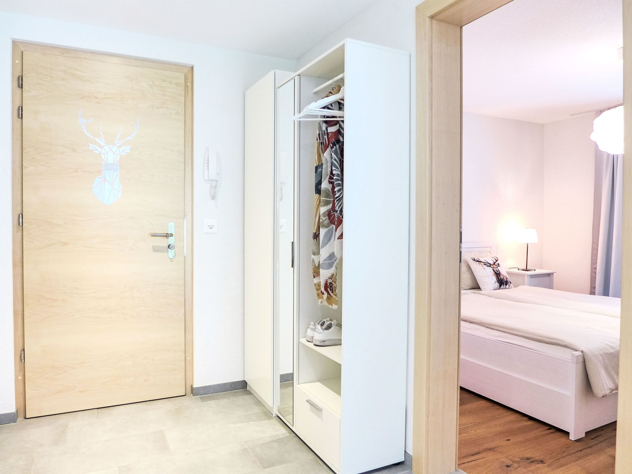 Habitación de hotel con una puerta de madera con diseño de cabeza de ciervo, un armario blanco con ropa colgada y una cama con cabecera blanca.