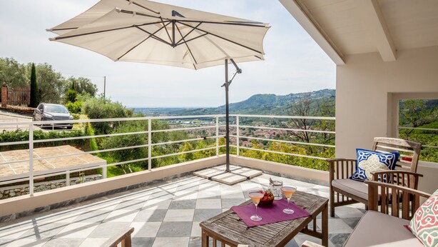 En balkon med et bord, stole og en parasol, med udsigt over et naturskønt landskab.