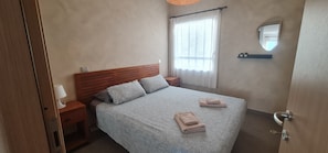 Une chambre à coucher avec une tête de lit en bois, un lit recouvert d’une literie à motifs, deux tables de chevet avec des lampes, une fenêtre avec des rideaux légers, un miroir fixé au mur et une porte avec un trou de serrure.