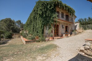 A stone house with a balcony, surrounded by a gravel driveway and greenery.