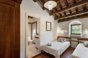 Ein Schlafzimmer mit zwei Betten, ein Badezimmer mit gefliestem Duschbereich und ein Fenster mit Tageslicht.