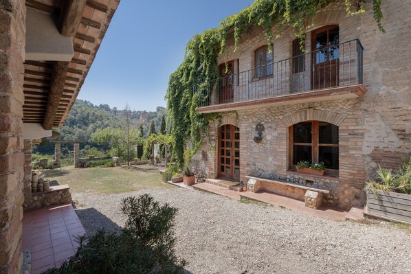 A stone building with a balcony, a gravel courtyard, and a view of a forested hillside.