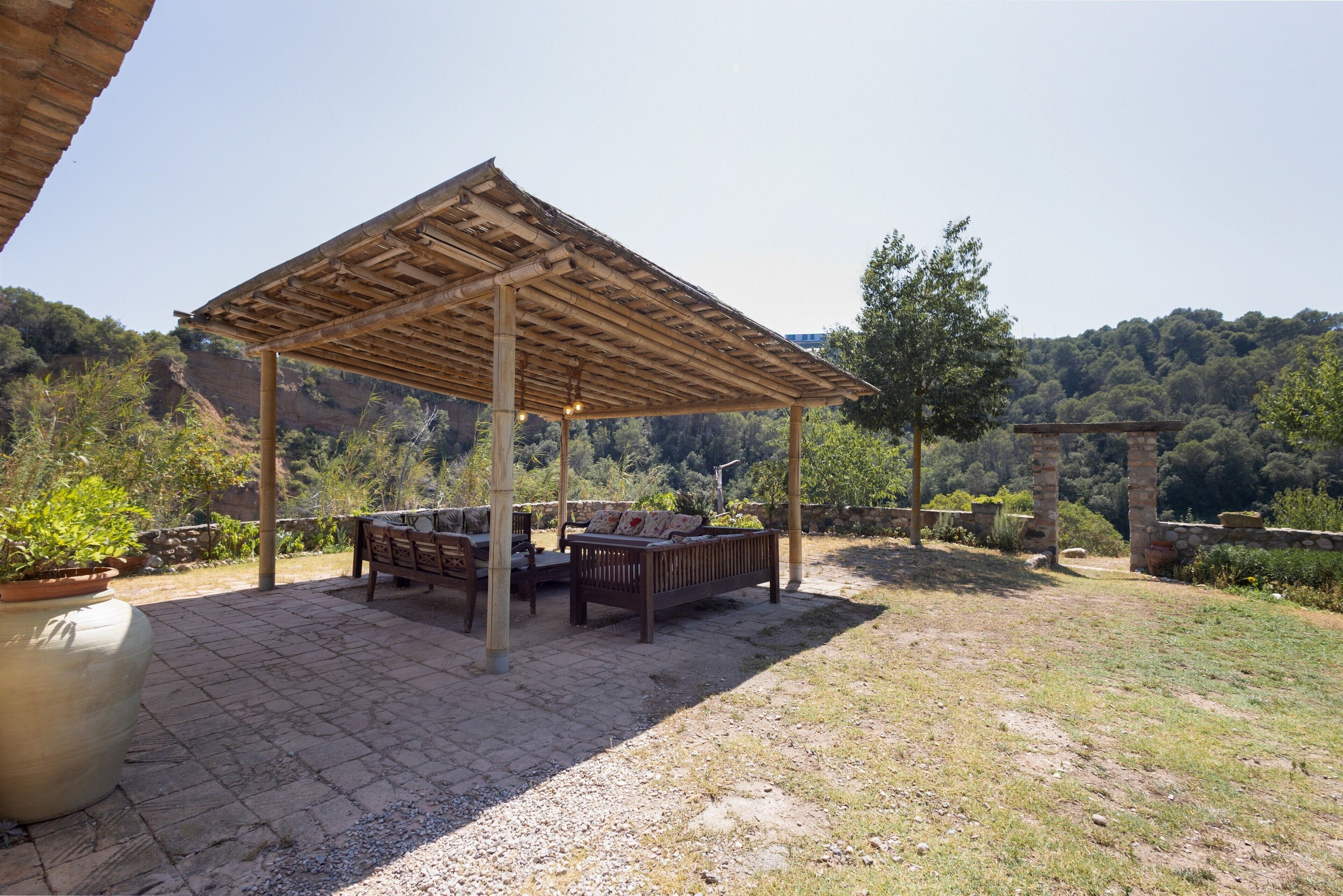 A covered outdoor seating area with a table and benches, surrounded by potted plants and a view of a mountainous landscape.