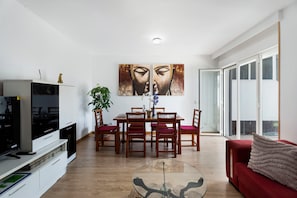 A modern living room with a flat-screen TV, dining table with chairs, a glass coffee table, and a red sofa.