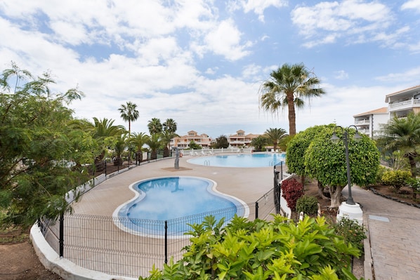 A swimming pool with a curved design, surrounded by a fence, palm trees, and a building with balconies in the background.