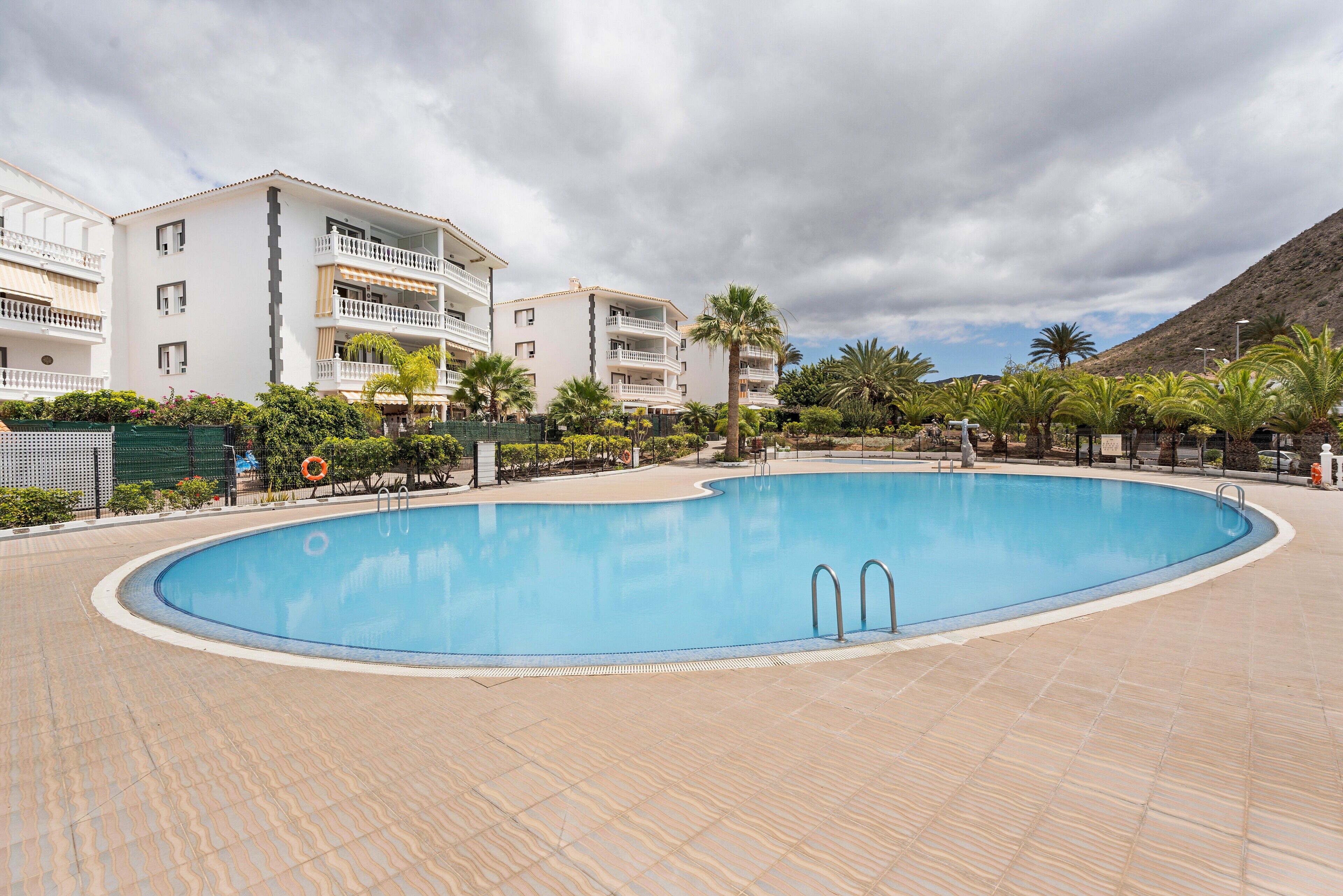 A swimming pool surrounded by a tiled deck, with a building featuring balconies and a mountainous backdrop.