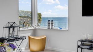 Quarto moderno com uma grande janela com vista para um corpo de água e vegetação. Há uma cadeira única, uma pequena mesa com objetos decorativos e uma estante com livros e um vaso.