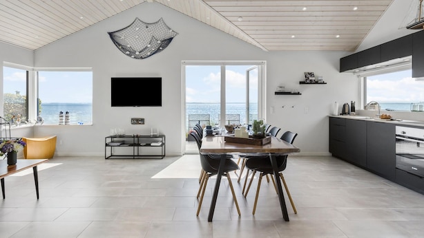 Uma sala de estar ampla com uma grande janela com vista para o mar, uma TV de tela plana na parede e uma cozinha moderna com armários escuros.