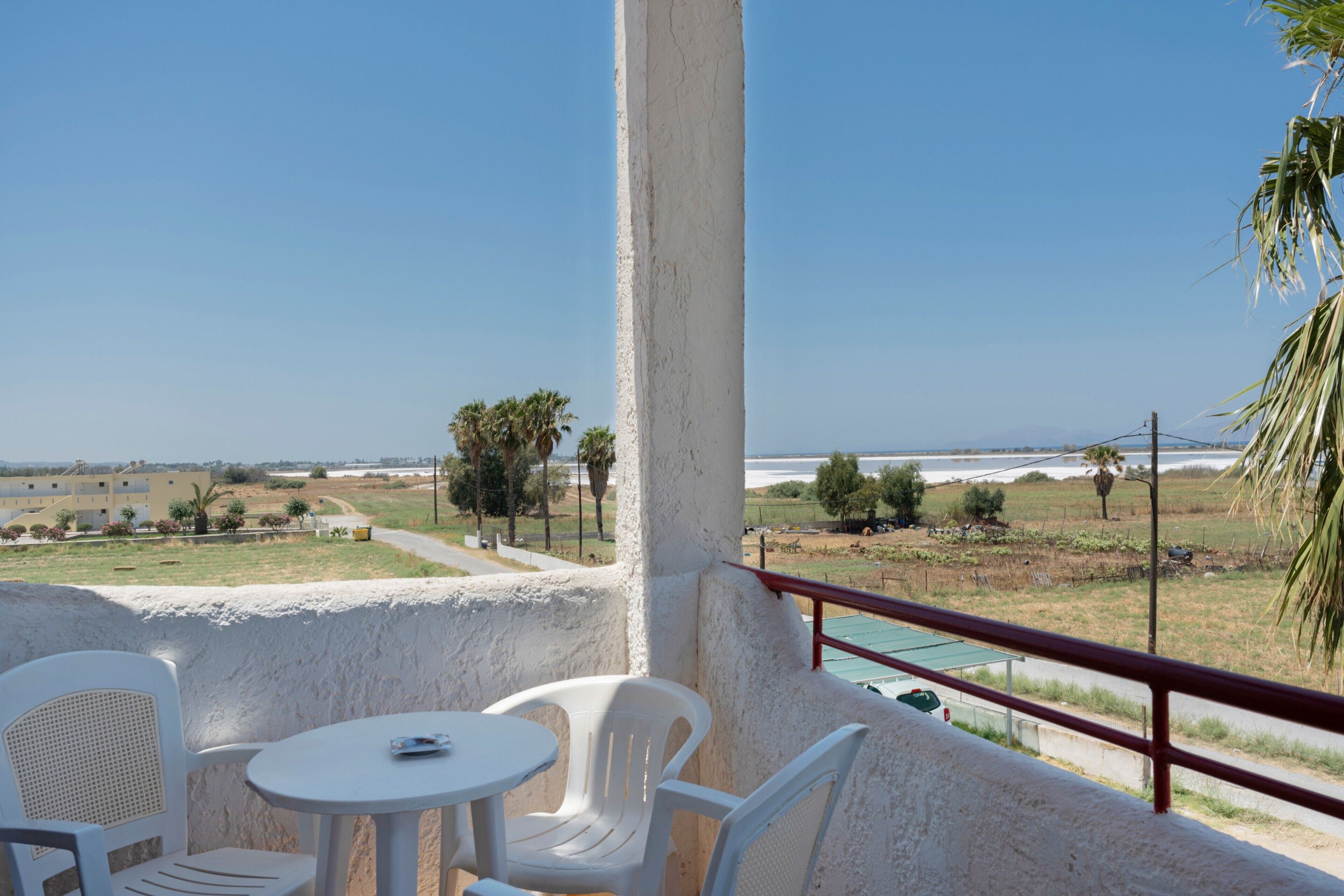 Un balcón con sillas de plástico blanco y una mesa pequeña, con vistas a un paisaje costero con edificios y palmeras.