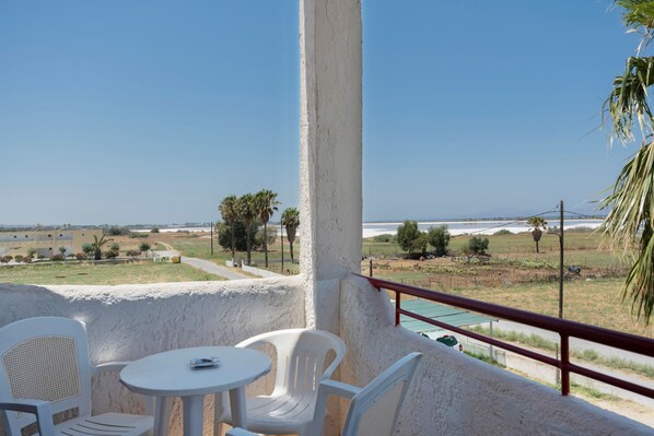Un balcon avec des chaises en plastique blanches et une petite table, donnant sur un paysage côtier avec des bâtiments et des palmiers.