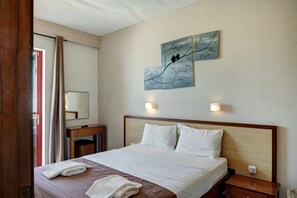 Habitación de hotel con cama, dos lámparas fijadas a la pared, una mesita de noche de madera, un aparador de madera y un cuadro de pájaros en una rama.