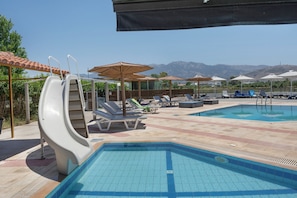 A swimming pool with slides, lounge chairs, umbrellas, and a mountainous backdrop. - Studio Apartment 'Golden Sun  8' with Sea View, Shared Pool and Wi-Fi (Tigaki 0)