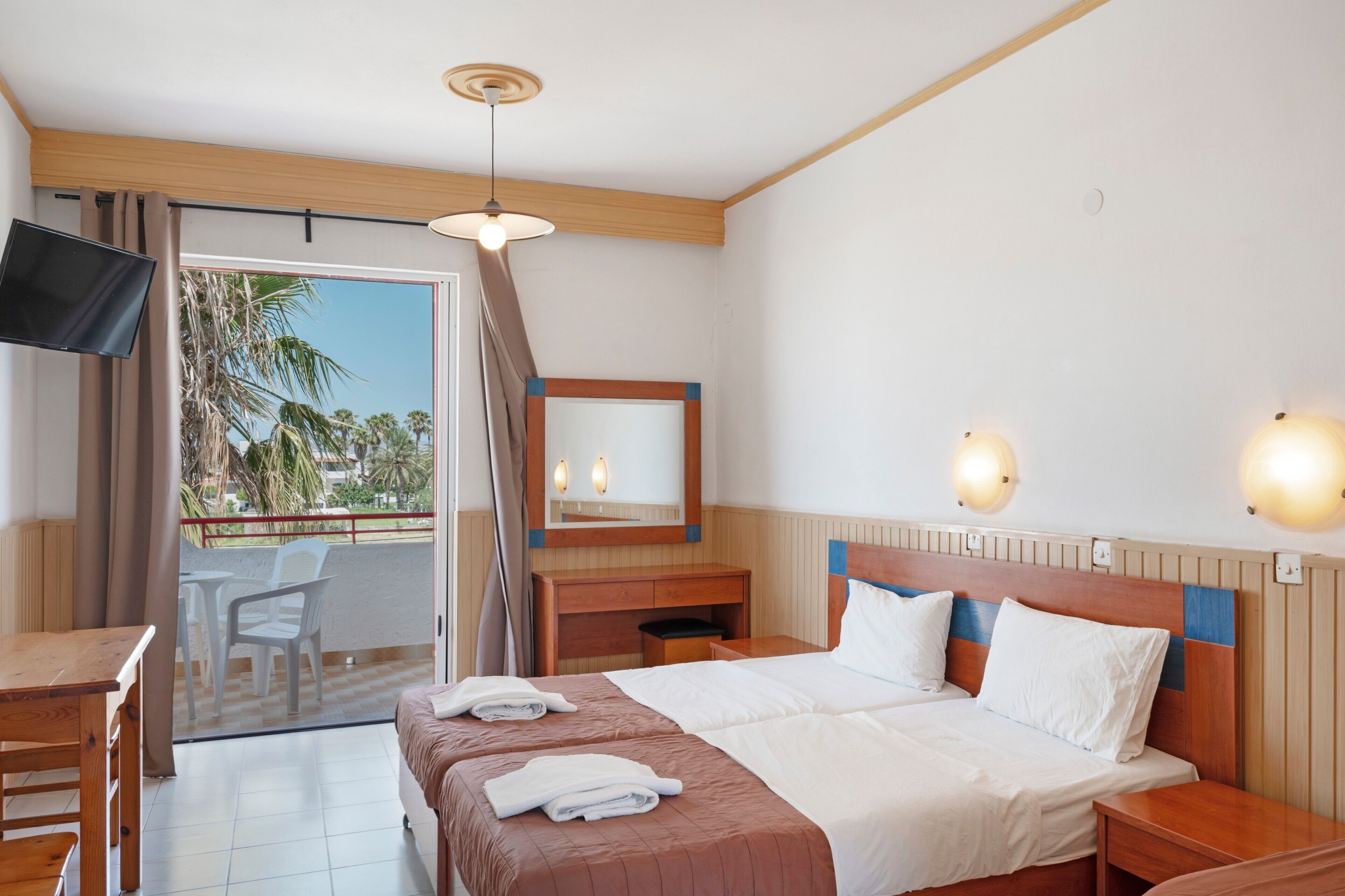 A hotel room with a bed, bedside tables, a desk, a TV, a balcony with chairs, and a view of palm trees.